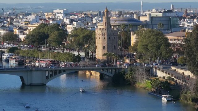 Aerial view of Seville, Andalusia shows Torre del Oro, an arched bridge, market tents, boats and a rower in late afternoon light, with locals and visitors moving