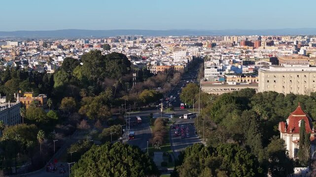 Aerial view shows Seville's Parque de Maria Luisa, a tree lined boulevard, buses and cars moving, pedestrians on shaded paths, wide framing, slow upward pan in daylight.