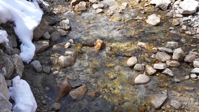 Mountain stream clear water flowing over rock, boulder and pebble on riverbed winter snowbank beside creek, sunlight reflections, alpine wilderness texture, cold tranquil scenery