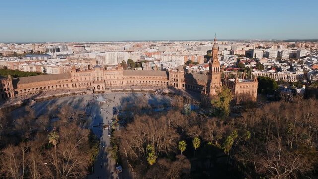 Aerial view of Plaza de Espana in Seville, Spain. Twin towers, central fountain, patterned bridges, and Parque de Maria Luisa trees in warm late afternoon light.