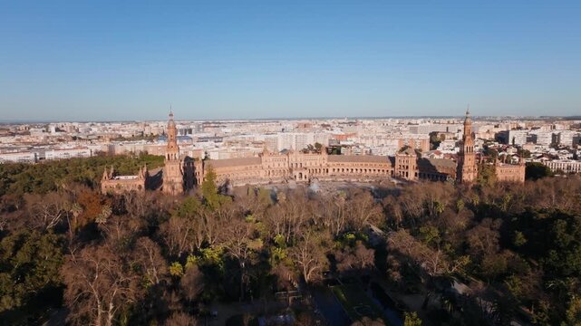Aerial view of Plaza de Espana in Seville as a drone pulls back. Twin towers, semicircular palace, ornate bridges, and pulsing fountains sit by Maria Luisa Park.