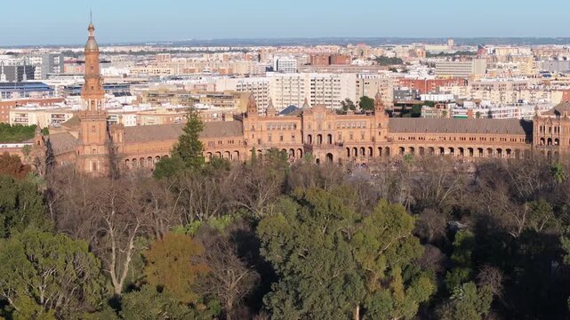 Drone pans from treetops to colonnades as visitors stroll. Twin towers, tiled bridges, and central fountain in warm late afternoon light in Seville, Spain.