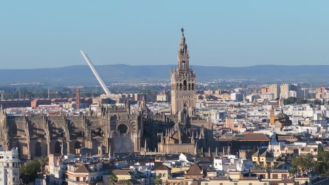 Aerial pan over central Seville, Andalusia, Spain. Giralda bell tower and Cathedral dominate as Calatrava bridge appears left, rooftops lead to distant hills.