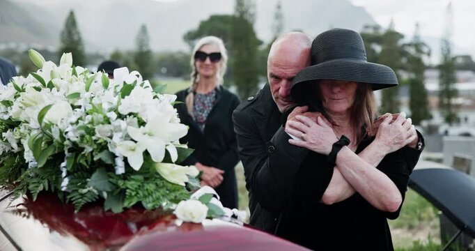 Coffin, flowers and senior couple at funeral, burial service or hug for comfort at cemetery. Man, elderly woman and white rose on casket for peace, sad and embrace to console while mourning death