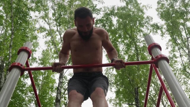 Low angle shot of a strong, shirtless man with a beard performing a difficult calisthenics exercise on a pull-up bar at an outdoor gym. He shows incredible strength and determination.