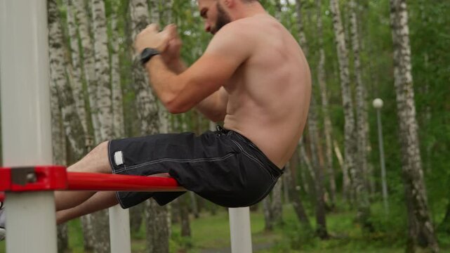 A shirtless athletic man with a beard performs abdominal crunches on parallel bars at an outdoor gym. He finishes the set and takes a break, breathing heavily.