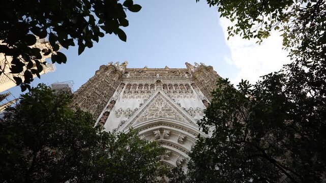 Low angle view of Seville Cathedral portal with tracery, statues, pointed arches, and orange trees. Giralda tower peeks left in late afternoon natural light.
