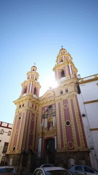 Baroque church in urban Spain rises above narrow street at midday as sun flares between towers, lighting statues, oculi, pilasters, and crosses, with cars and pedestrians present.
