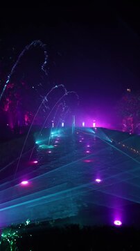 Night in Spain shows an illuminated fountain with arcing jets, a long reflecting pool, hedges, and lanterns, with magenta and cyan light and laser like beams.