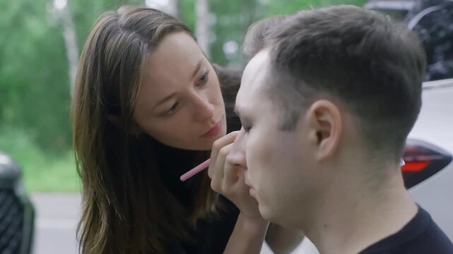 A female makeup artist carefully applies dark eyeshadow or eyeliner to a young man's eye with a brush. The scene is outdoors, suggesting preparation for a film shoot or photoshoot.