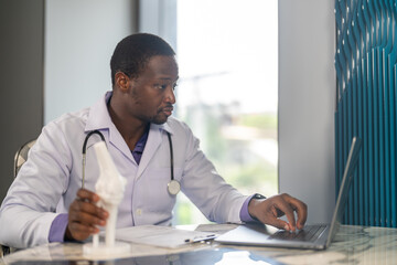 Fototapeta premium African American male physical therapist doctor looking at laptop holding anatomical knee joint model in rehabilitation center, Orthopedic medical clinic, healthcare and physiotherapy concept