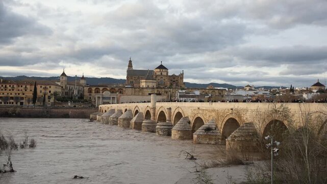 Crowds walk across stone arches as Mezquita Catedral dome and bell tower rise behind historic walls. Late afternoon light and clouds cast warm sandstone tones.