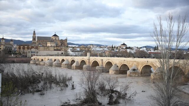 The Roman Bridge spans a swollen Guadalquivir in Cordoba as pedestrians move, muddy currents swirl around cutwaters, and late light highlights warm sandstone tones.