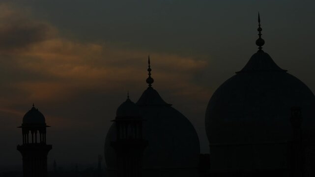 Time-lapse footage of a mosque silhouette with domes and minarets set against a colorful sky during sunrise or sunset. The dark architectural outline contrasts beautifully with soft hues.