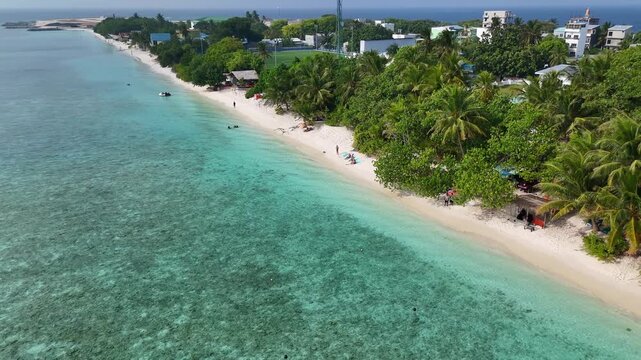 Beautiful drone shot flying over a tropical beach paradise in the Maldives. People sunbathe on white sand next to vibrant blue ocean and lush green trees.