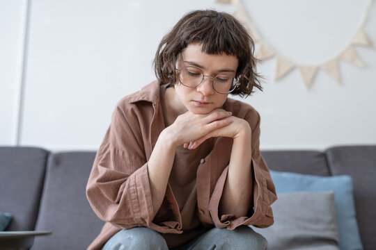 Sad upset teenage girl sitting on sofa, looking down with withdrawn expression, resting chin on hands. Teen uncertainty, social isolation, low self-confidence, introversion and emotional discomfort.