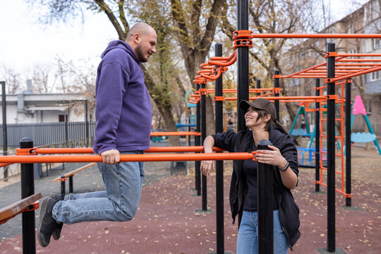 Man performing dips on parallel bars while talking to a woman at an outdoor fitness park during daytime.