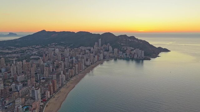 Stunning aerial footage showing the beautiful city of benidorm during a golden sunrise. The tranquil mediterranean sea and skyline are visible