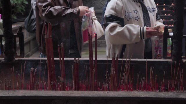 Close-up static shot of visitors holding a small wooden divination box over a large incense burner to absorb rising incense smoke during a traditional Buddhist ritual at Luohan Temple in Chongqing
