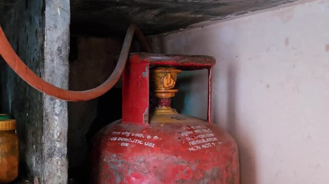 A weathered orange LPG cylinder sits in a dim alcove, its yellow regulator and hose ready for use. Amidst the 2026 supply crisis and price hikes, this essential Indian kitchen staple home cooking.