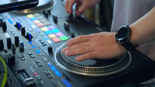 Close-up of DJ hands scratching vinyl on professional turntable controller with colorful RGB pads, faders and knobs in studio or club setting