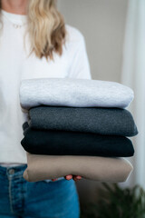 Fototapeta premium Vertical lifestyle image of blonde woman in jeans and white shirt holding neutral sweatshirts in clean bedroom, small business owner or laundry concept
