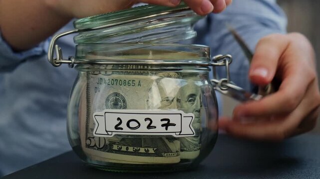 Female hands take money from Glass jar full of American currency dollars cash banknote with text 2027 year. Preparation saving money. Moderate consumption and economy. Collecting money
