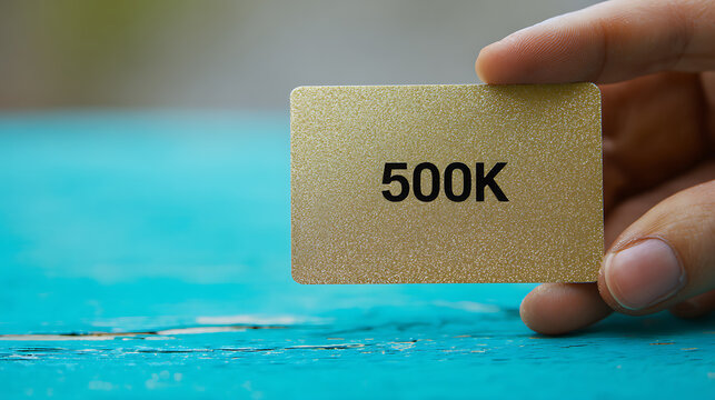 Human hand hold a shiny golden card showing "500K" across a vibrant blue table, signifying a milestone or achievement