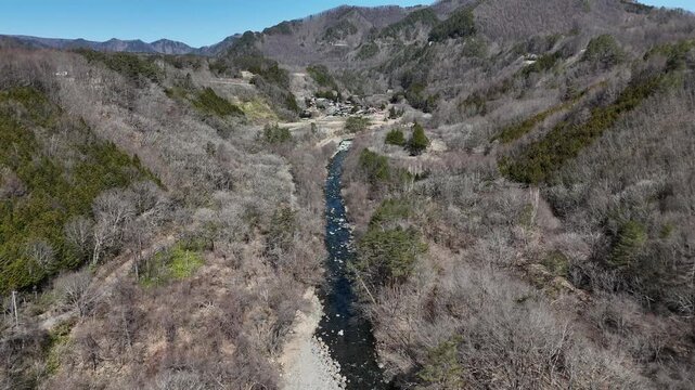 Aerial view shows a river winding through a valley. Trees are bare, signaling late autumn or early spring. Foliage is sparse, patches of green break the brown