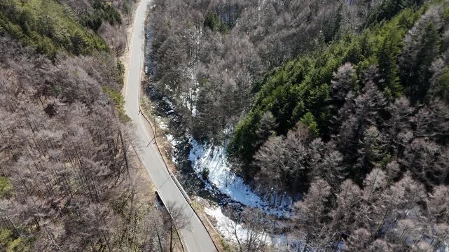 Aerial view shows winding road through snowy forested valley. Road cuts through bare trees and evergreens. Snow patches linger near riverbed below. Vehicle moves slowly along the narrow path