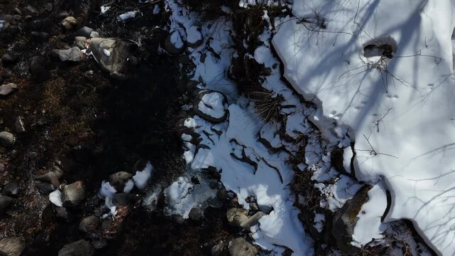 Snow melts near rocks and dirt. Riverbed shows winter's retreat. Patches of ice linger beside snow. Sunlight casts tree shadows. Nature's quiet transformation. Perfect for seasonal change scenes