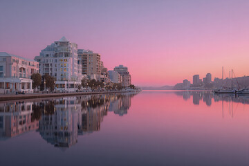 Fototapeta premium Sunrise waterfront cityscape with soft pink sky tones reflecting on calm water, modern buildings and boats create peaceful urban atmosphere at dawn