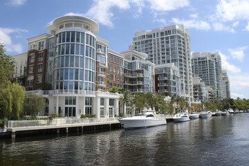 Obraz premium Modern waterfront condominium complex with yachts docked along calm river under bright blue sky, surrounded by lush greenery and palm trees creating serene atmosphere