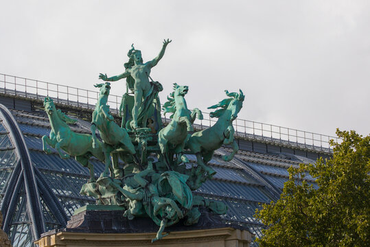 Sculpted chariot surges skyward above a glass iron roof in Paris. Bronze horses rear as a figure reaches outward with dramatic motion. Green patina glows against pale sky over the vast hall.