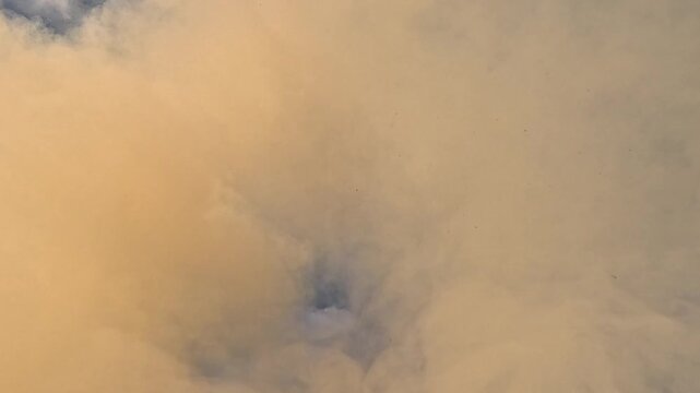 A pale beige smoke cloud expands across the blue sky, then billows, softens and drifts outward, forming a dusty atmospheric background with changing density and slow natural motion.