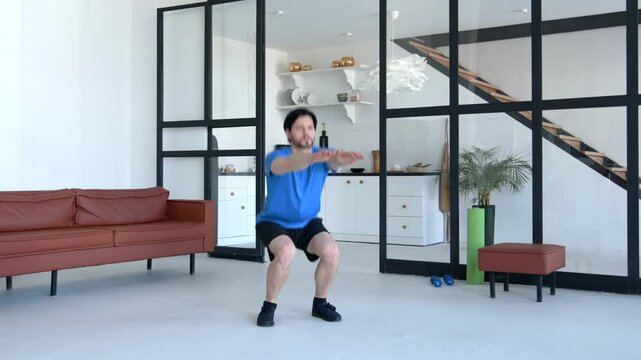 Motivated active fitness man in sportswear practicing squats exercise, improving core strength and mobility, building stringer leg muscles while working out in domestic room.