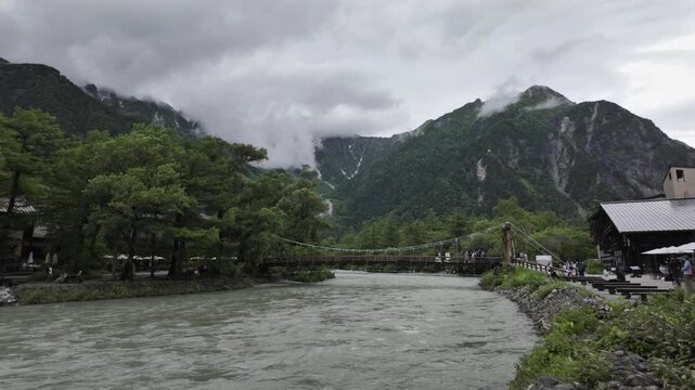 A suspension bridge over the turbulent river, the famous Kappa Bridge in Kamikochi. 
Cloudy weather with tall mountains in the distance.