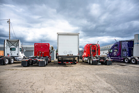 Back view of the different big rig semi trucks and semi trailers standing in row on industrial parking lot with stormy sky