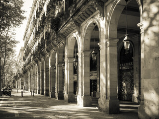 Sepia-toned arcade of a neoclassical building with stone arches, hanging lanterns and tree-lined boulevard. Barcelona, Spain. © davidrbcelta