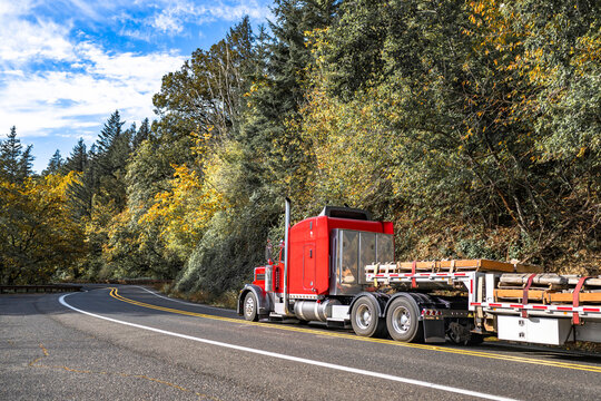 Bright red classic big rig semi truck with stainless steel boxes on the back wall transporting lumber cargo on step deck semi trailer running on the winding autumn road with forest on the side