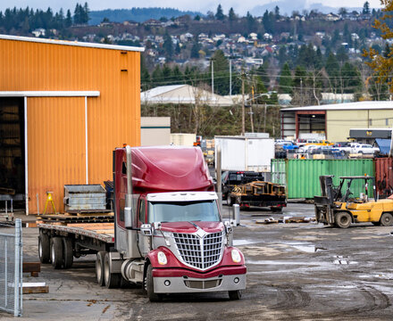 Burgundy big rig semi truck with flat bed semi trailer loading cargo standing on the industrial fabrication lot