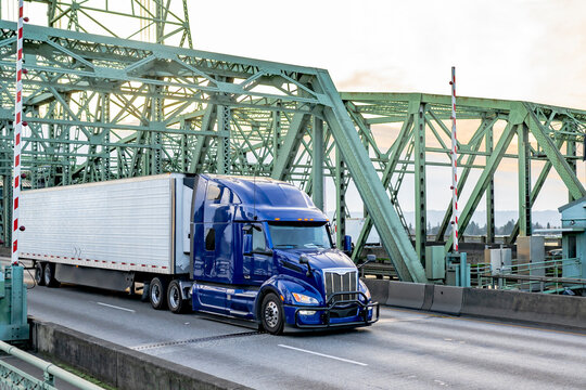 Dark blue big rig semi truck tractor transporting cargo in refrigerator semi trailer running on the truss bridge at twilight time