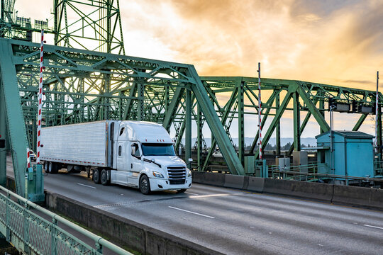 High cab white big rig semi truck transporting cargo in refrigerator semi trailer running on the truss bridge at twilight time