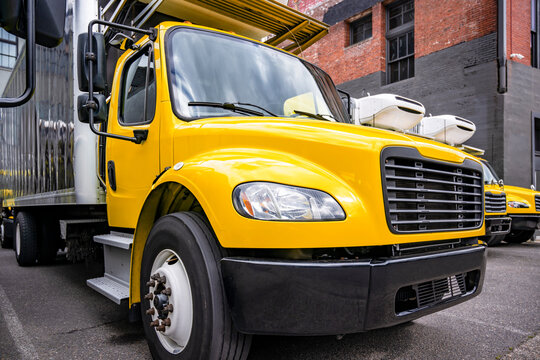 Middle duty yellow day cab rig semi truck with refrigerated box trailer standing in row with another semi trucks on industrial parking lot waiting for the next load