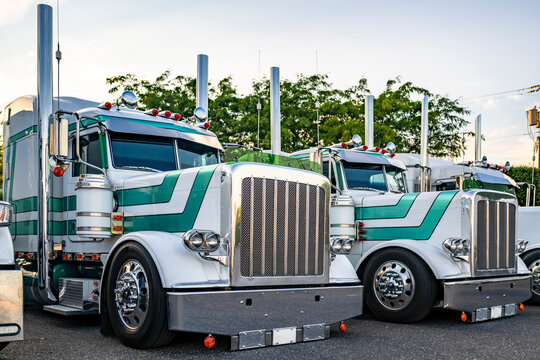 Old style powerful classic American big rig semi trucks tractors with chrome accessories and high exhaust pipes standing in row on truck stop parking lot at twilight time