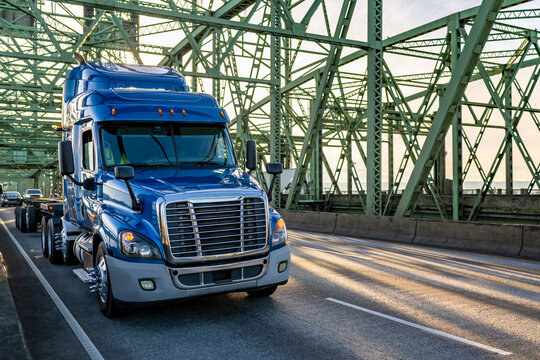 Popular low cab profile blue big rig semi truck with flat bed semi trailer running on the truss green bridge at sunset gold hour
