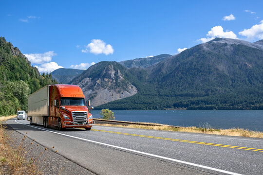 Orange big rig semi truck with grille guard transporting cargo in dry van semi trailer running on the highway road along the Columbia River in Columbia River Gorge area