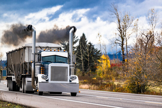 Powerful old style black big rig semi truck tractor with black smoke from the exhaust pipes polluting the environment transporting cargo in bulk semi trailer running on the autumn road