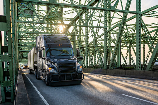 Stylish black big rig semi truck transporting fastened cargo on step deck semi trailer running on the truss draw bridge in sunshine