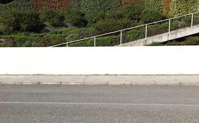 Naklejka premium White plaster fence with a staircase, vegetation and multicolored hedge on behind. Concrete sidewalk and street in front. Background for copy space.